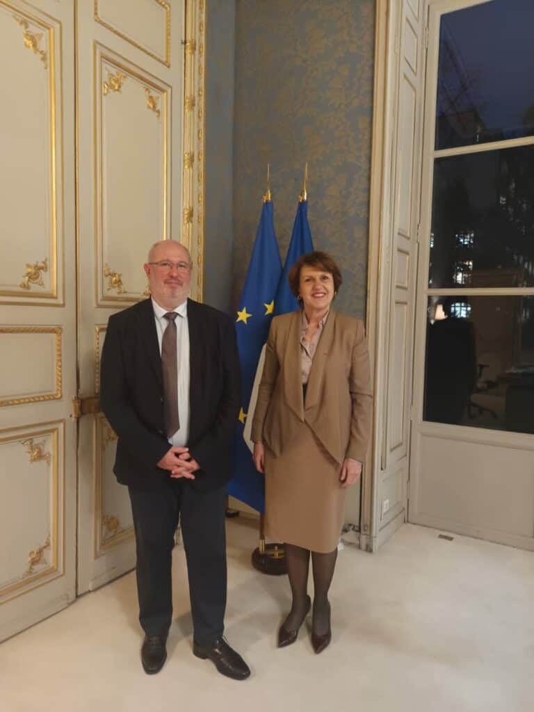 Une rencontre officielle entre Eric Sargiacomo, député européen, et une représentante lors d’un événement politique, avec les drapeaux de l’Union Européenne en arrière-plan dans un cadre élégant et formel.