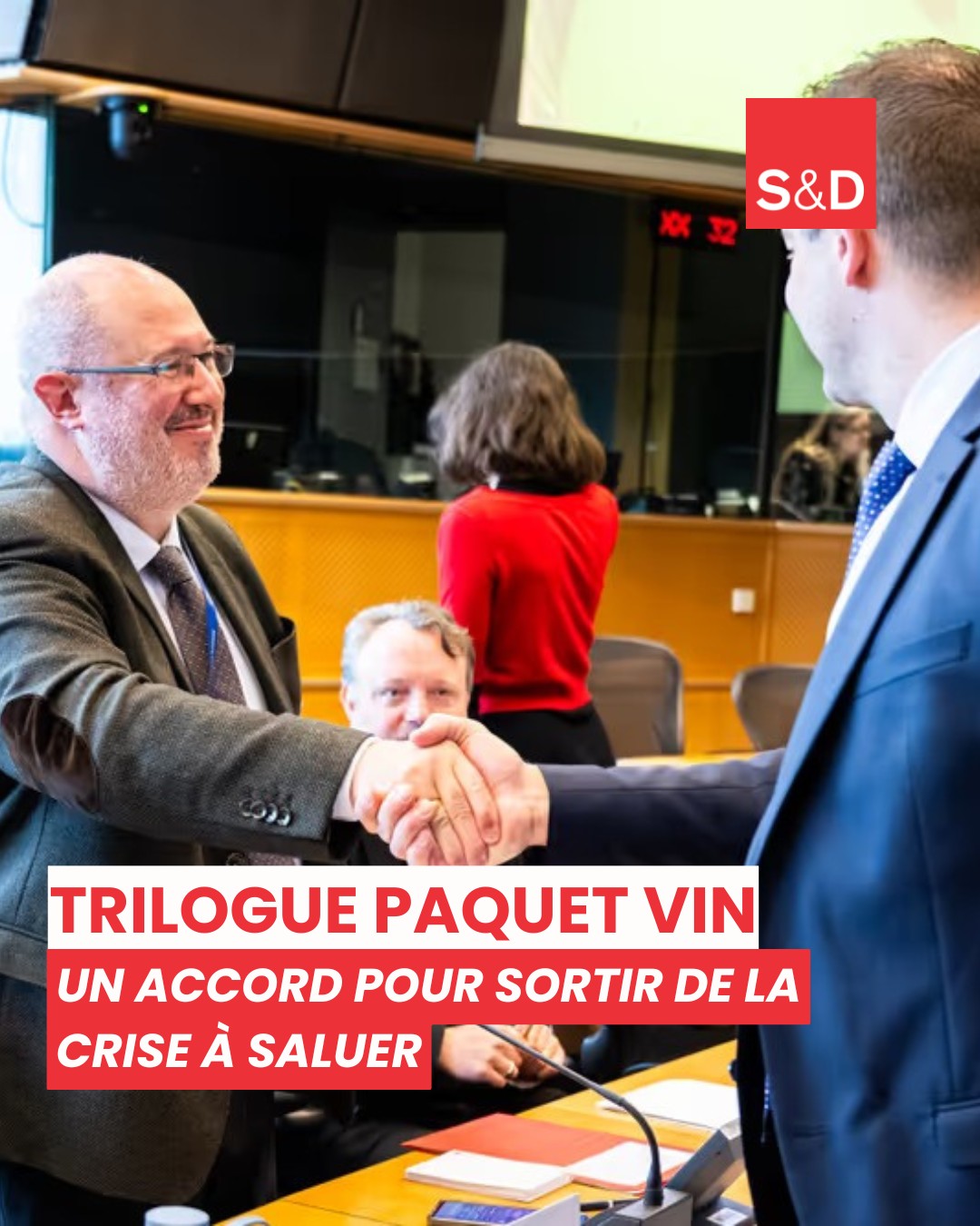 1. Portrait d'un homme politique européen serrant la main d'un autre lors d'une réunion officielle, avec des collègues en arrière-plan, dans une salle de conférence moderne.